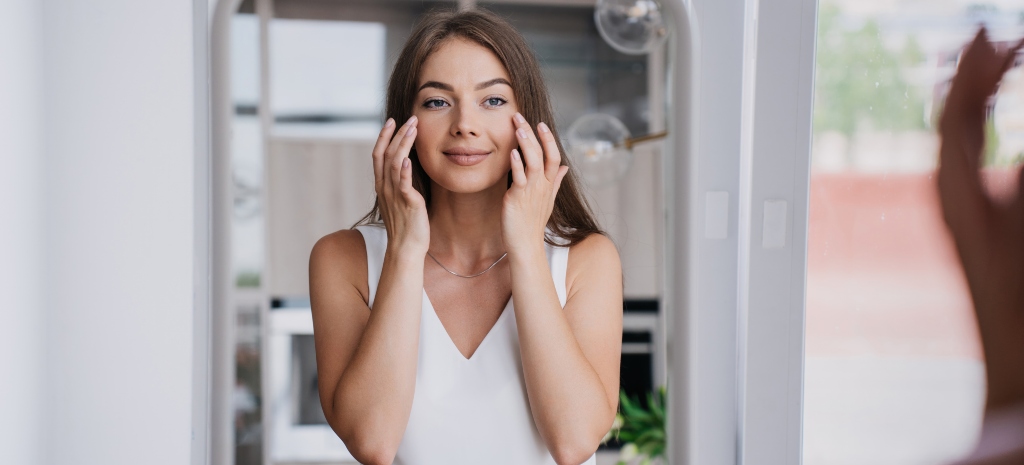 Beautiful woman massaging her face in the mirror after facelift procedure, which we offer at our plastic surgery centers in Los Angeles, San Francisco, and Orange County.