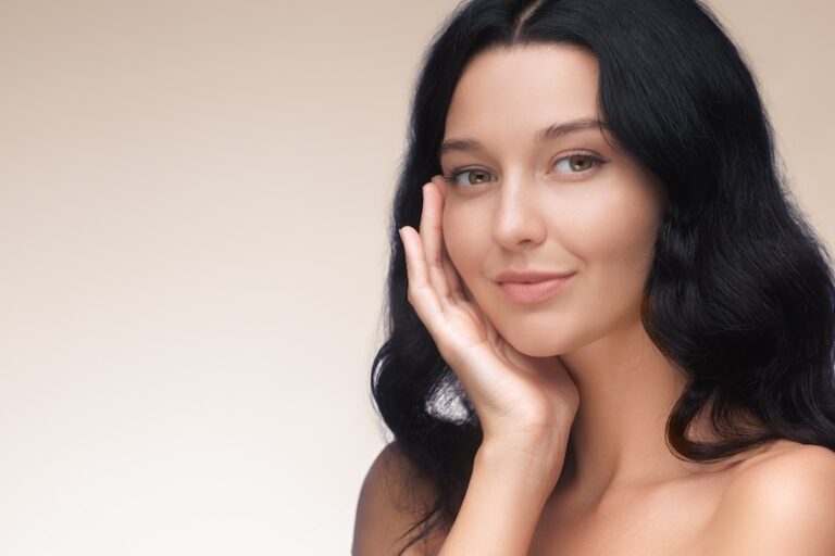 Happy young adult woman touching her face with hand on cheek after facial harmony surgery on beige studio background.