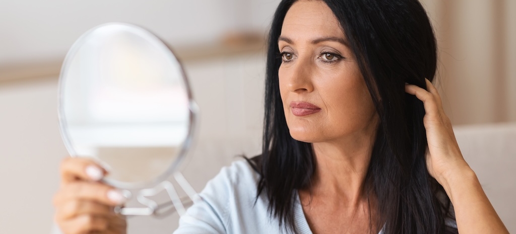 Portrait of beautiful middle-aged brunette lady holding mirror and looking at her reflection, touching her hair with hand. Confident adult woman thinking about dermal fillers