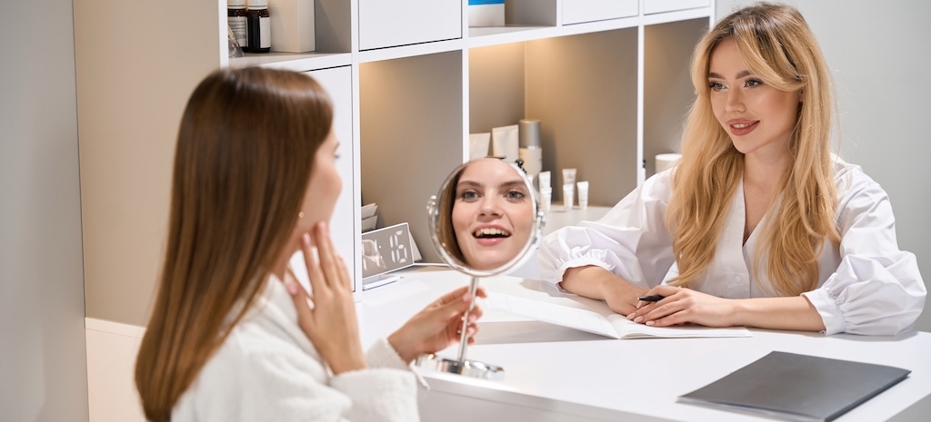 Female aesthetic technician looking at young smiling woman looking at herself in mirror during consultation at desk.