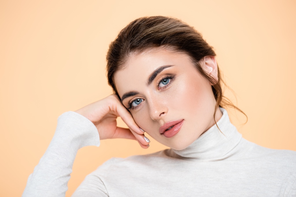 stylish woman in turtleneck looking at camera while touching face isolated on peach