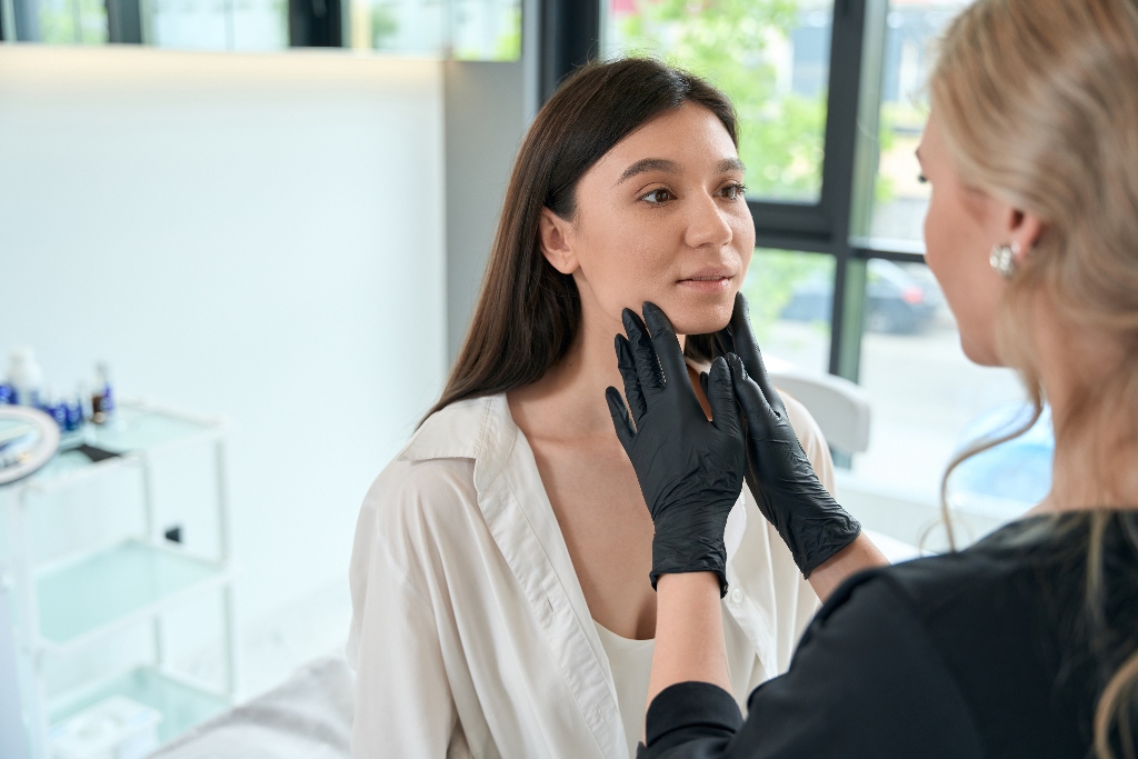 Female in medical coat and protective gloves standing near client and looking at her face assessing options for minimally invasive anti-aging options