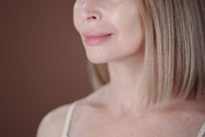 Senior woman smiling subtly with side view, showing wrinkles and smooth skin with blonde straight hair looking serene and content without makeup against plain background