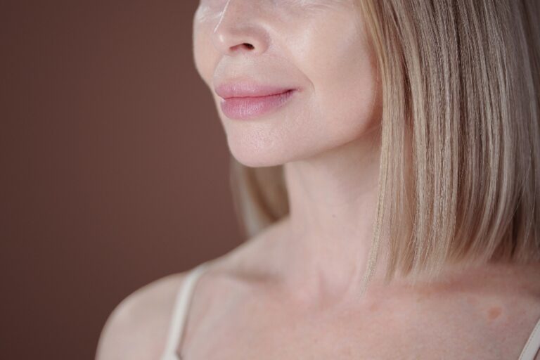 Senior woman smiling subtly with side view, showing wrinkles and smooth skin with blonde straight hair looking serene and content without makeup against plain background