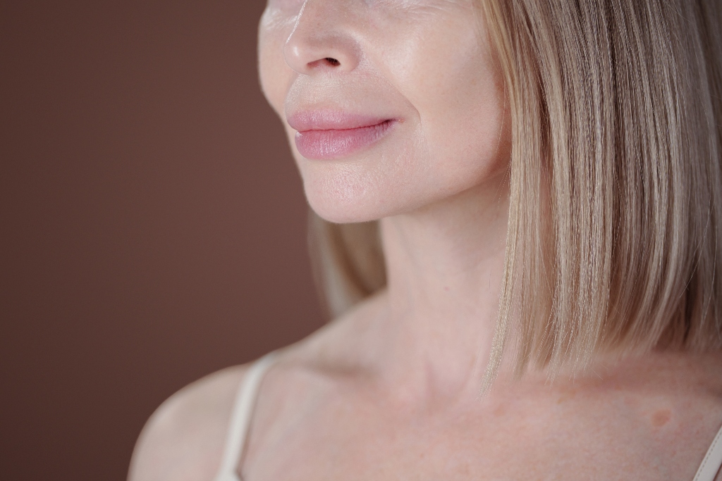 Senior woman smiling subtly with side view, showing wrinkles and smooth skin with blonde straight hair looking serene and content without makeup against plain background